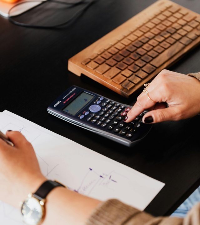From above crop anonymous female accountant working with calculator and writing down notes on paper while sitting at table with computer keyboard